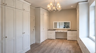 Fitted dressing room with painted floor-to-ceiling wardrobes, a dressing table and a window seat by C H Joinery Solutions